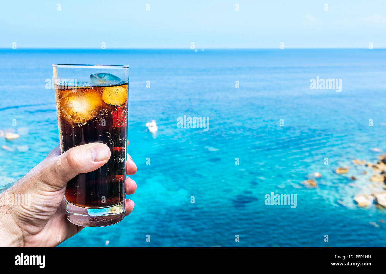 Mano azienda bicchiere con ghiaccio-raffreddata cola contro il blu del cielo e del mare Foto Stock