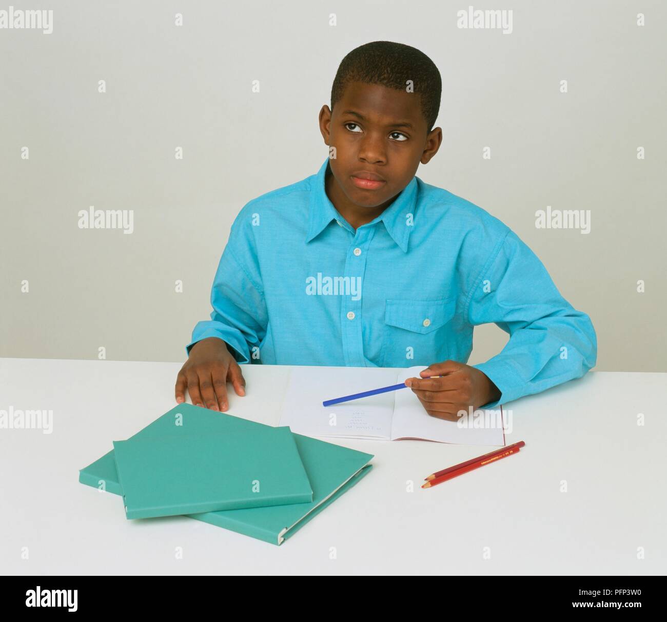 Ragazzo in maglietta blu seduto alla scrivania con carta e penna, guardando lontano Foto Stock