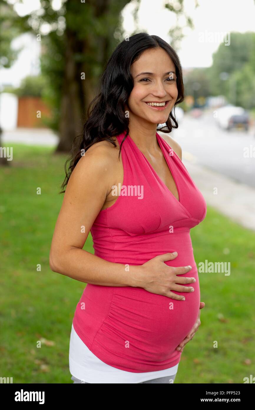 Donna incinta modeling rosa top senza maniche e le mani sul suo ventre, sorridente Foto Stock