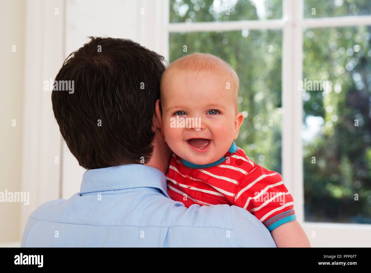Padre tenendo premuto 8 mese vecchio baby boy che è sempre sorridente e guardando sopra la sua spalla Foto Stock