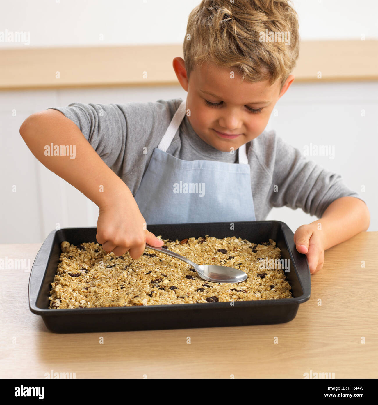 Ragazzo appiattimento muesli nel vassoio da forno con il dorso del cucchiaio, rendendo il muesli bar, 4 anni Foto Stock