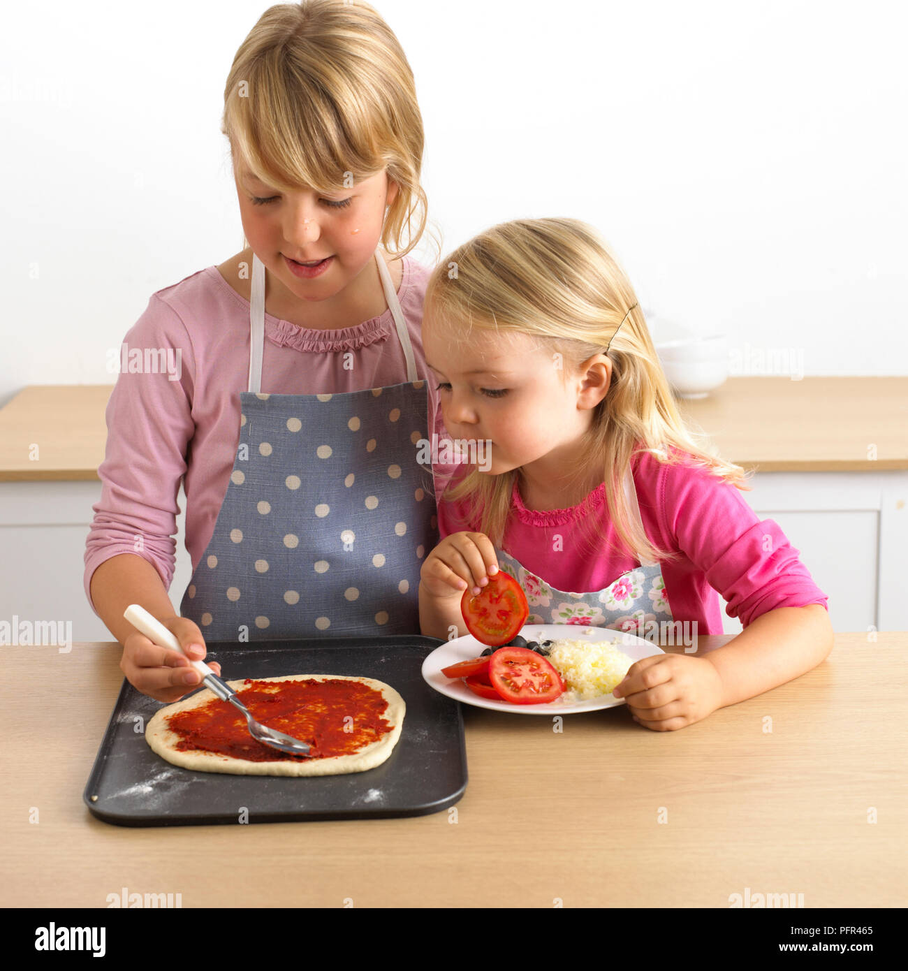 Due ragazze mettendo crema sulla pizza cruda base, 7 e 3 anni Foto Stock