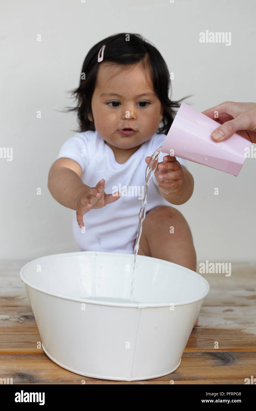 Baby girl (11 mesi) guardando come donna versa acqua dalla tazza in un recipiente Foto Stock