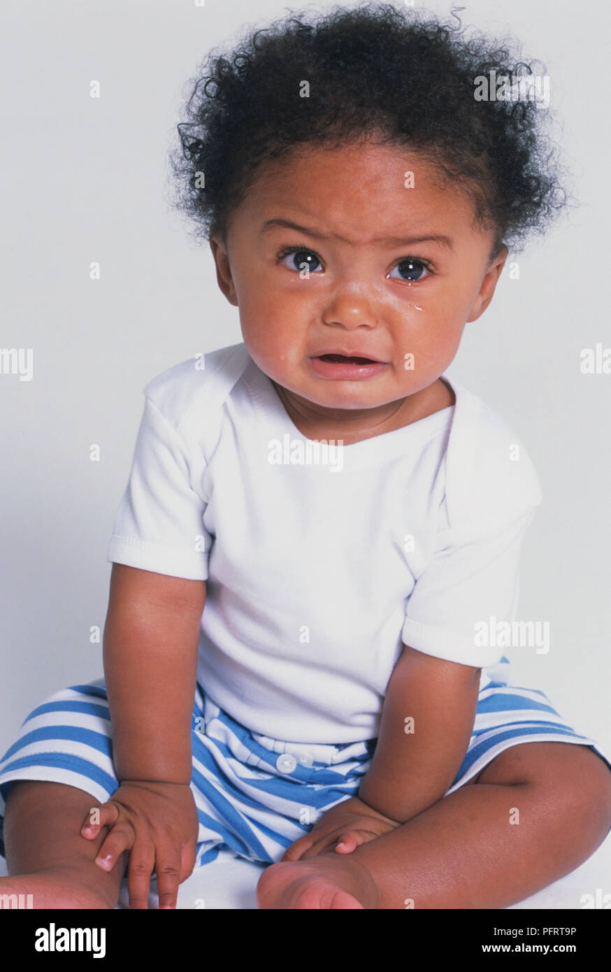 Baby boy (8,5 mesi) in pantaloncini a strisce seduto sul pavimento e gridando, vista frontale Foto Stock