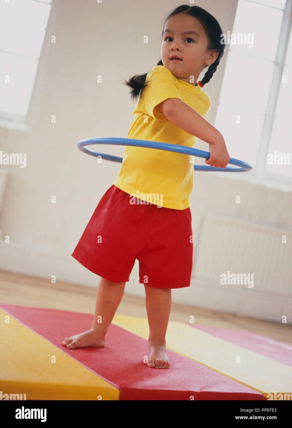 Il Toddler indossando giallo brillante, T-shirt e pantaloncini rossi in piedi sul tappeto a strisce preparazione di spin cerchio di plastica intorno alla vita Foto Stock
