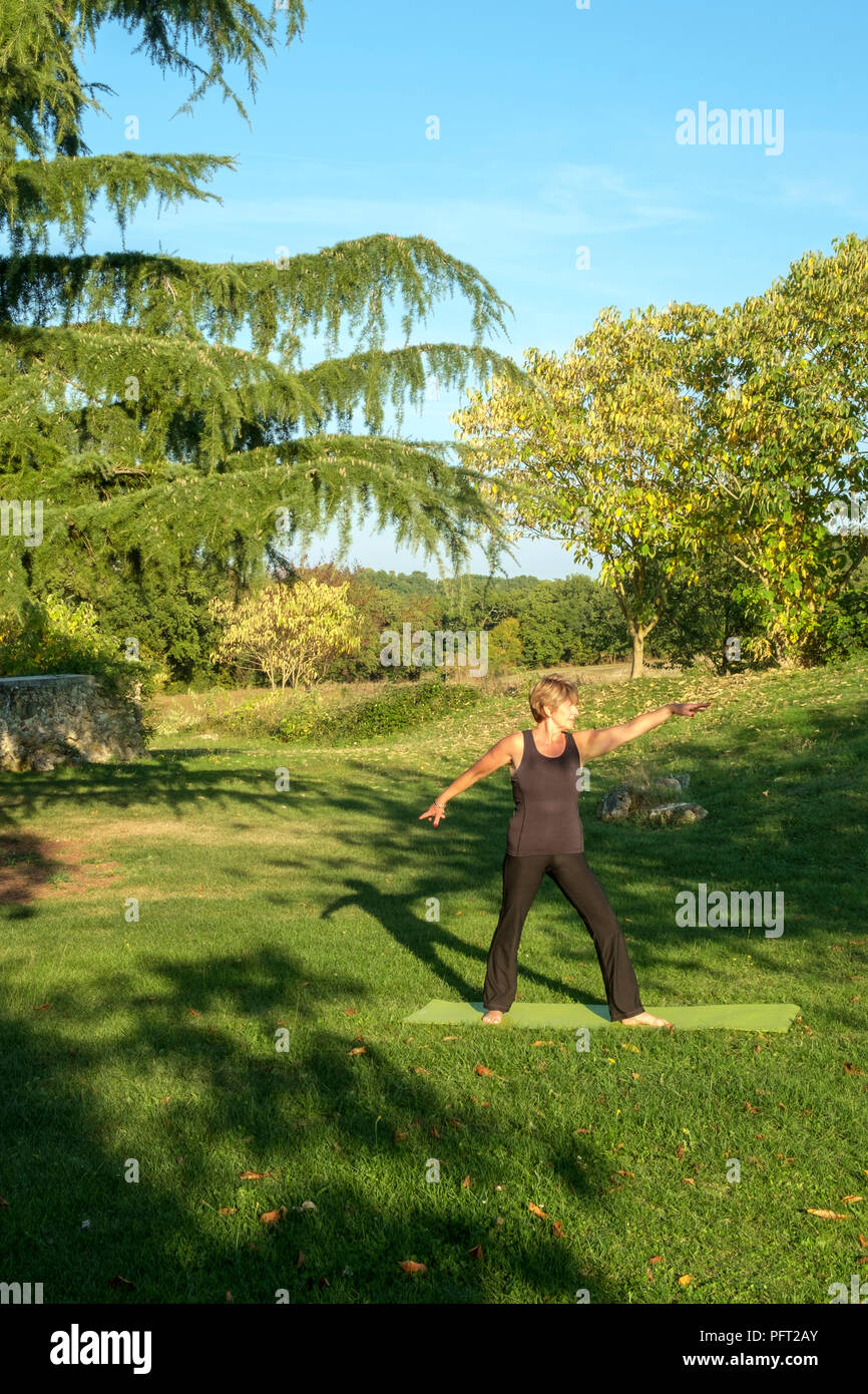Circondato dalla campagna idilliaca campagna francese una donna matura la sua prassi posizioni di yoga in autunno bello e mite sole. La posa del guerriero. Foto Stock