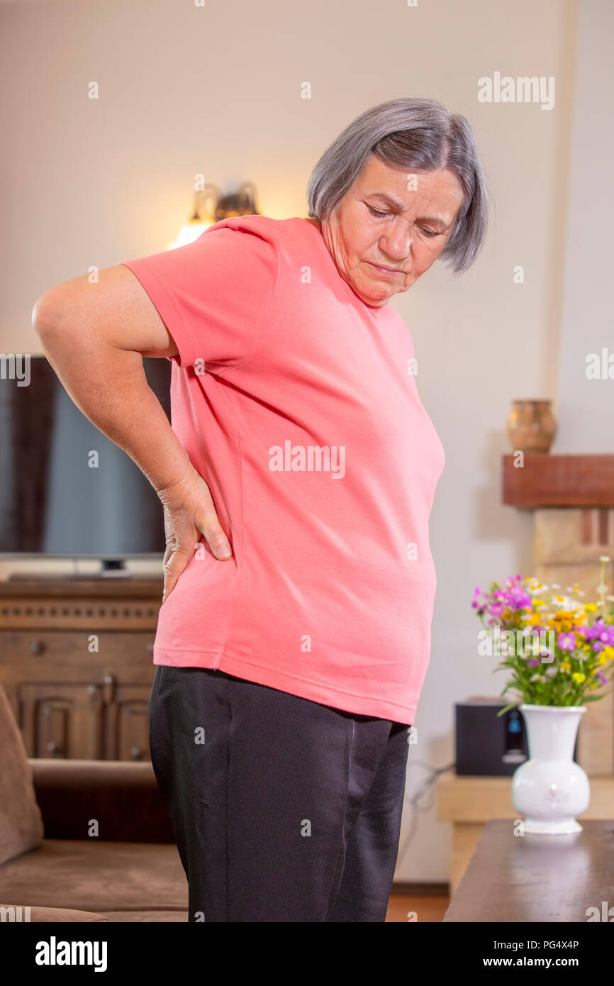 Donna matura che soffrono di mal di schiena a casa. Massaggio schiena inferiore con la mano, sensazione di esaurimento, in piedi nel salotto. Foto Stock