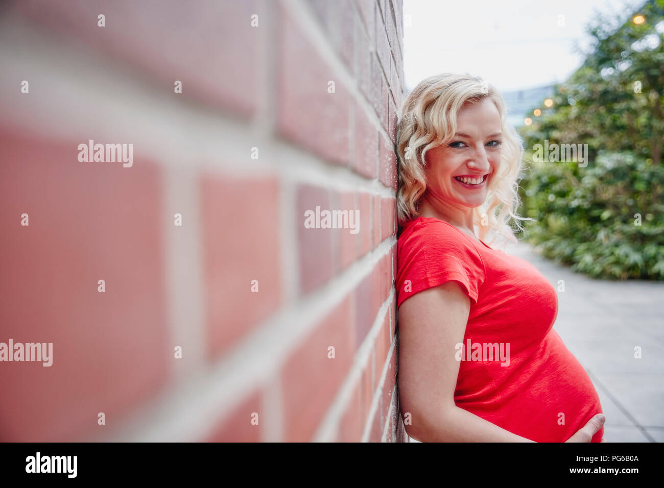Ritratto di sorridere donna incinta all'aperto appoggiata contro un muro di mattoni Foto Stock