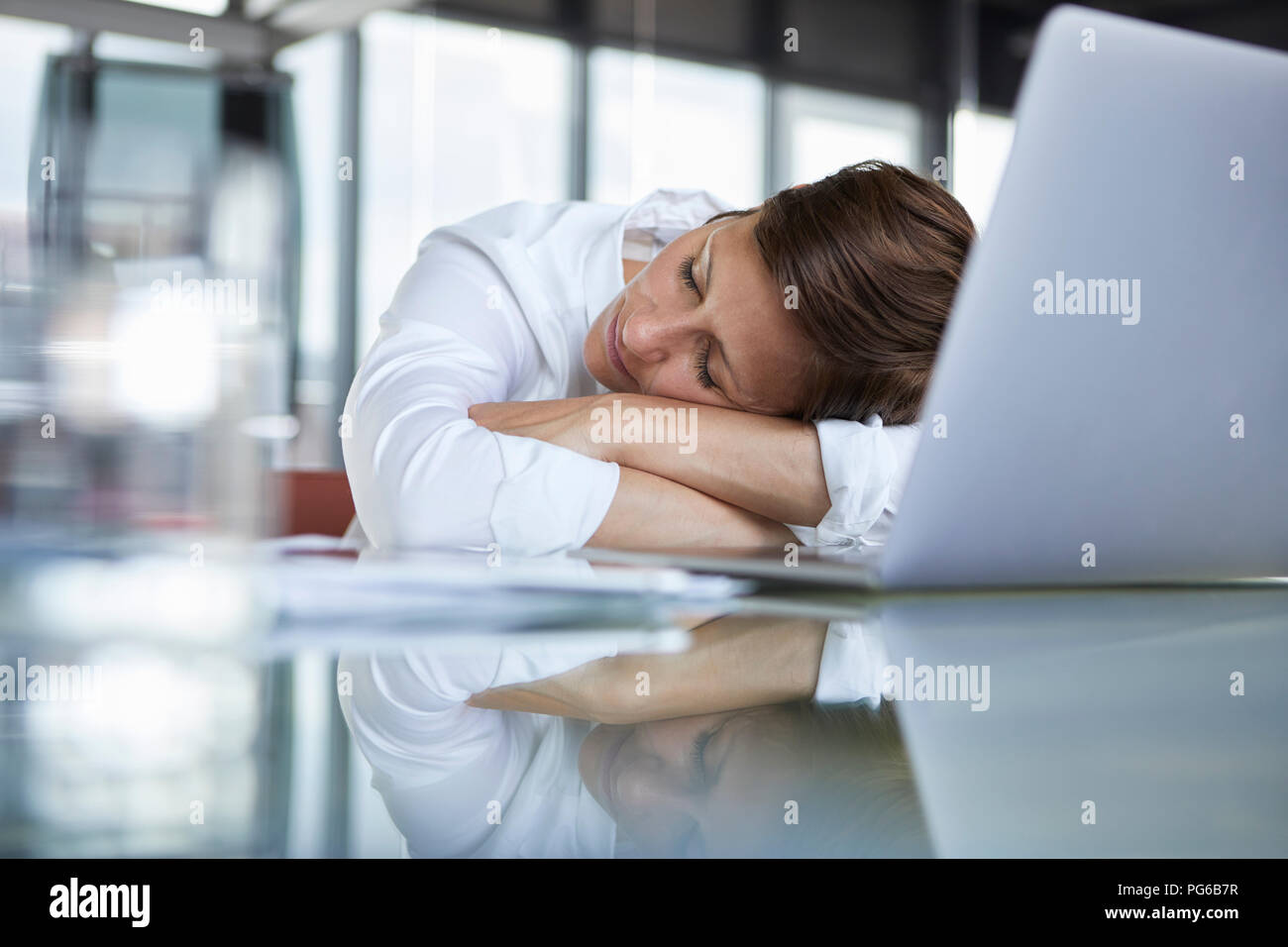 Imprenditrice con gli occhi chiusi che giace sul tavolo di vetro in ufficio nella parte anteriore del computer portatile Foto Stock