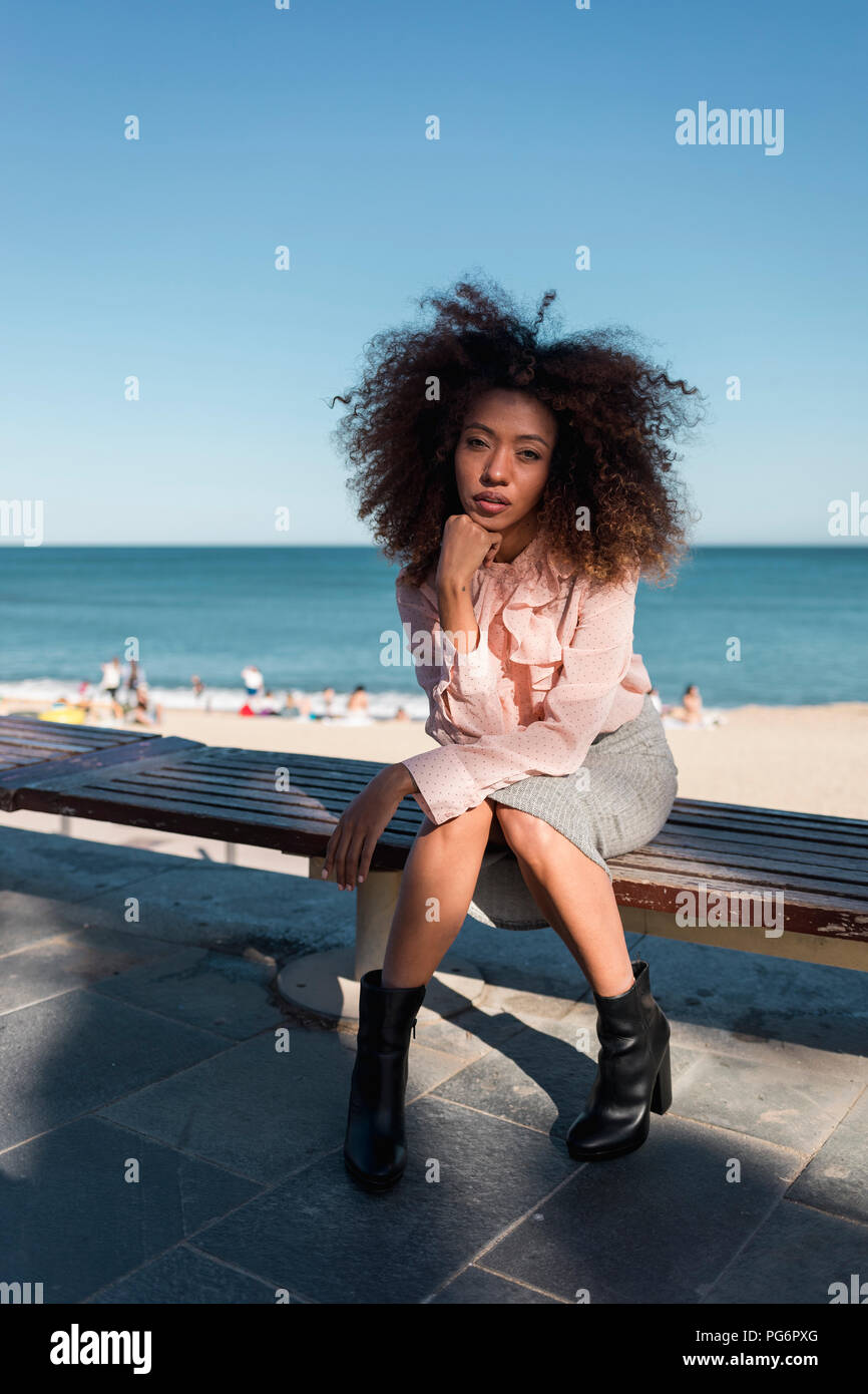 Ritratto di giovane e bella donna con taglio di capelli afro seduta su una panchina in spiaggia Foto Stock