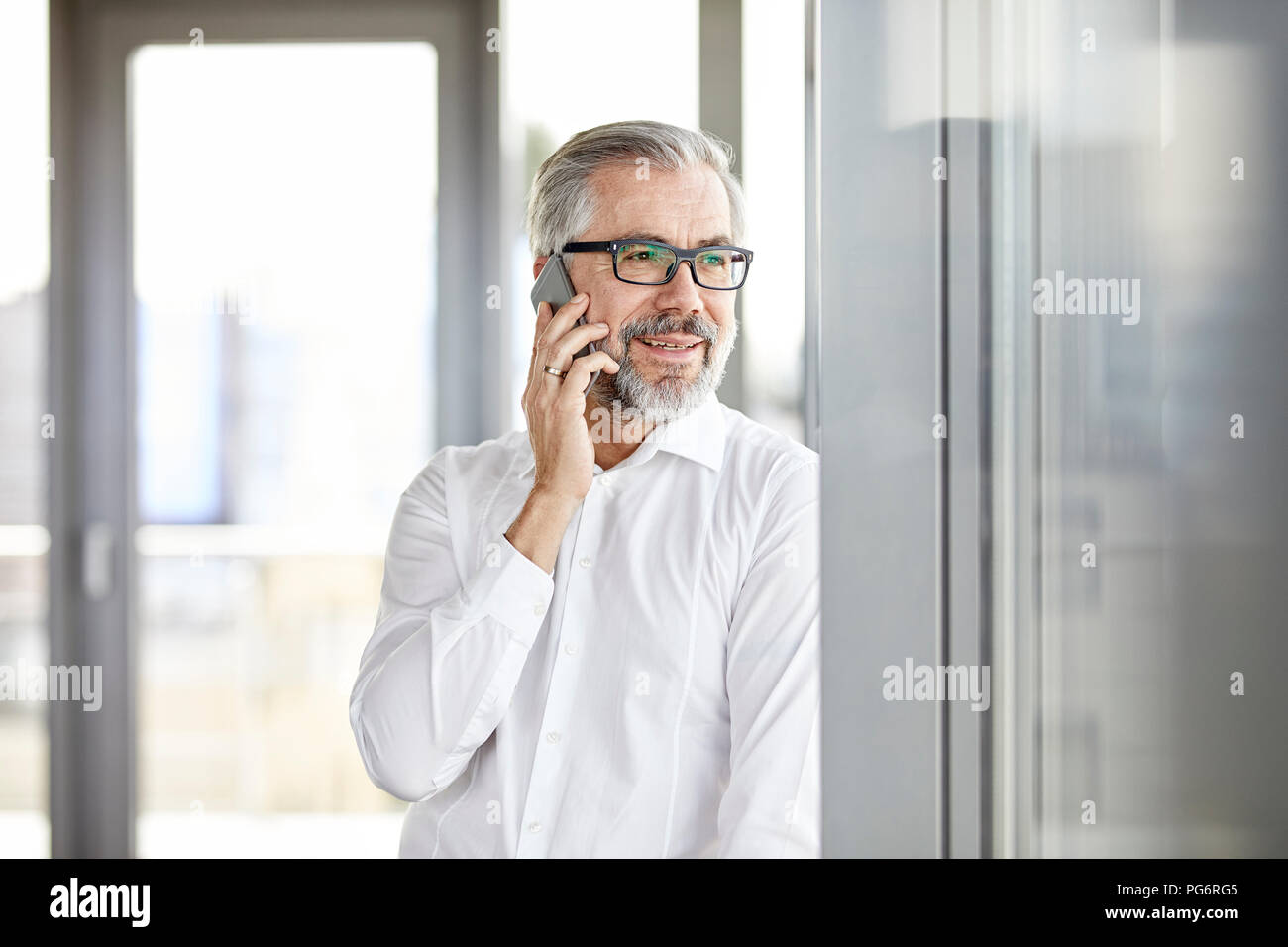 Imprenditore sorridente parlando al cellulare in corrispondenza della finestra Foto Stock