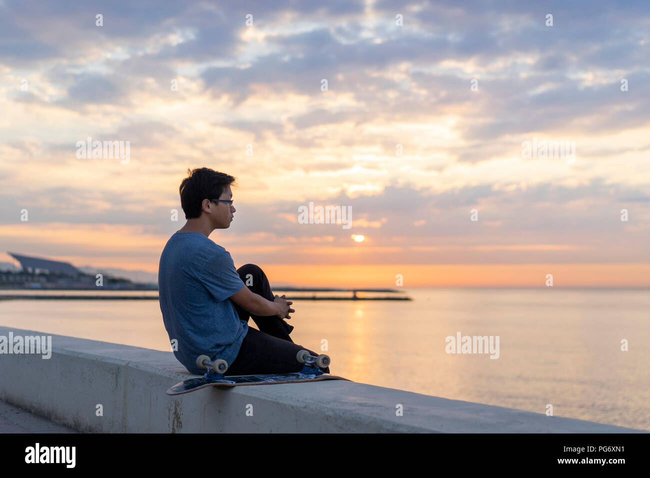 Giovane uomo cinese con lo skateboard seduti sulla parete in spiaggia Foto Stock