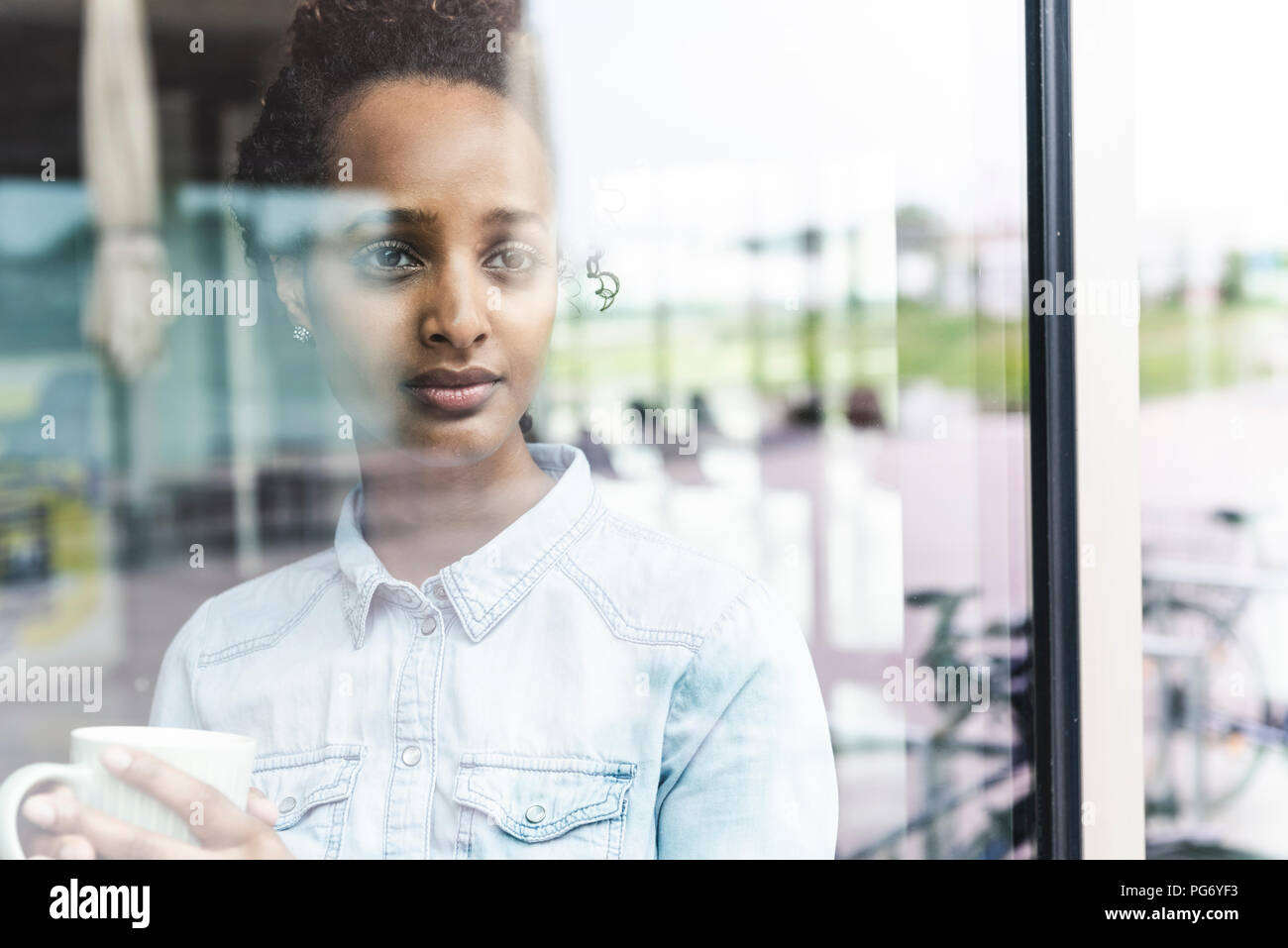 Ritratto di giovane imprenditrice guardando fuori della finestra, tenendo tazza da caffè Foto Stock