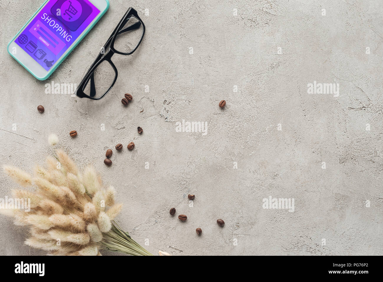 Vista dall'alto di smartphone con shopping app sullo schermo con occhiali, schizzi di caffè in grani e lagurus ovatus bouquet su una superficie in calcestruzzo Foto Stock