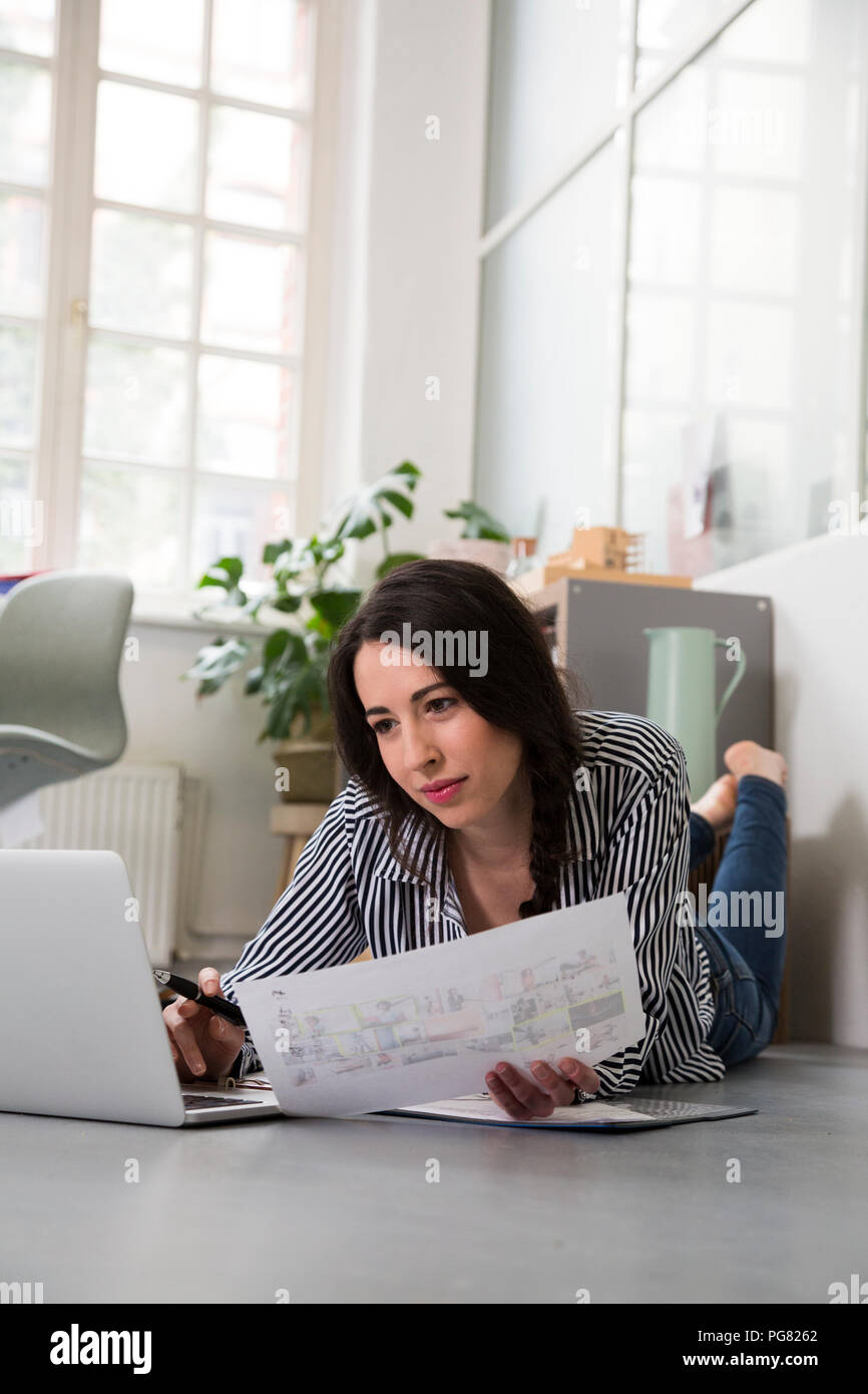 Donna informale con foglio di carta e laptop sdraiato sul pavimento in office Foto Stock