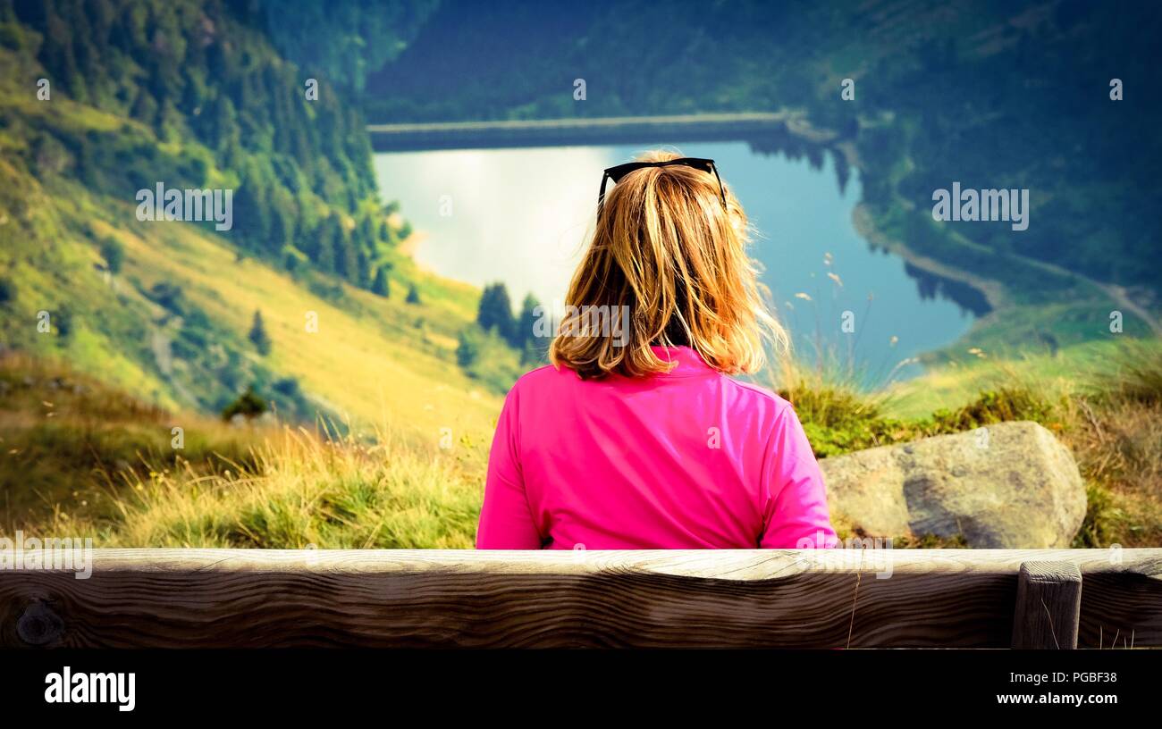 Una donna bionda seduto da solo in un banco in montagna. Ella è guardando verso il basso su una valle con un lago. Gusto Vintage colori Foto Stock