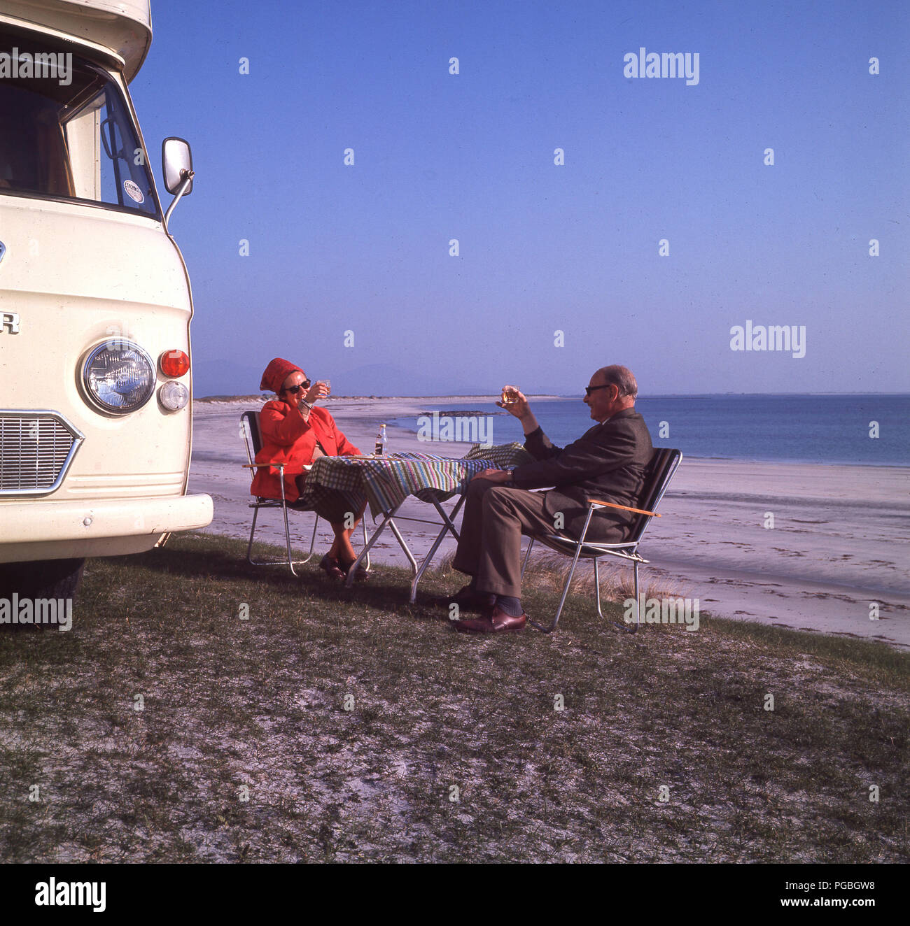 anni '70, grazie! Una coppia in pensione alzano un bicchiere per la salute di ciascuno degli altri, come si siedono fuori del loro camper Commer sulla costa accanto a una spiaggia di sabbia sull'isola di Harris negli altopiani scozzesi, Scozia, Regno Unito. Foto Stock