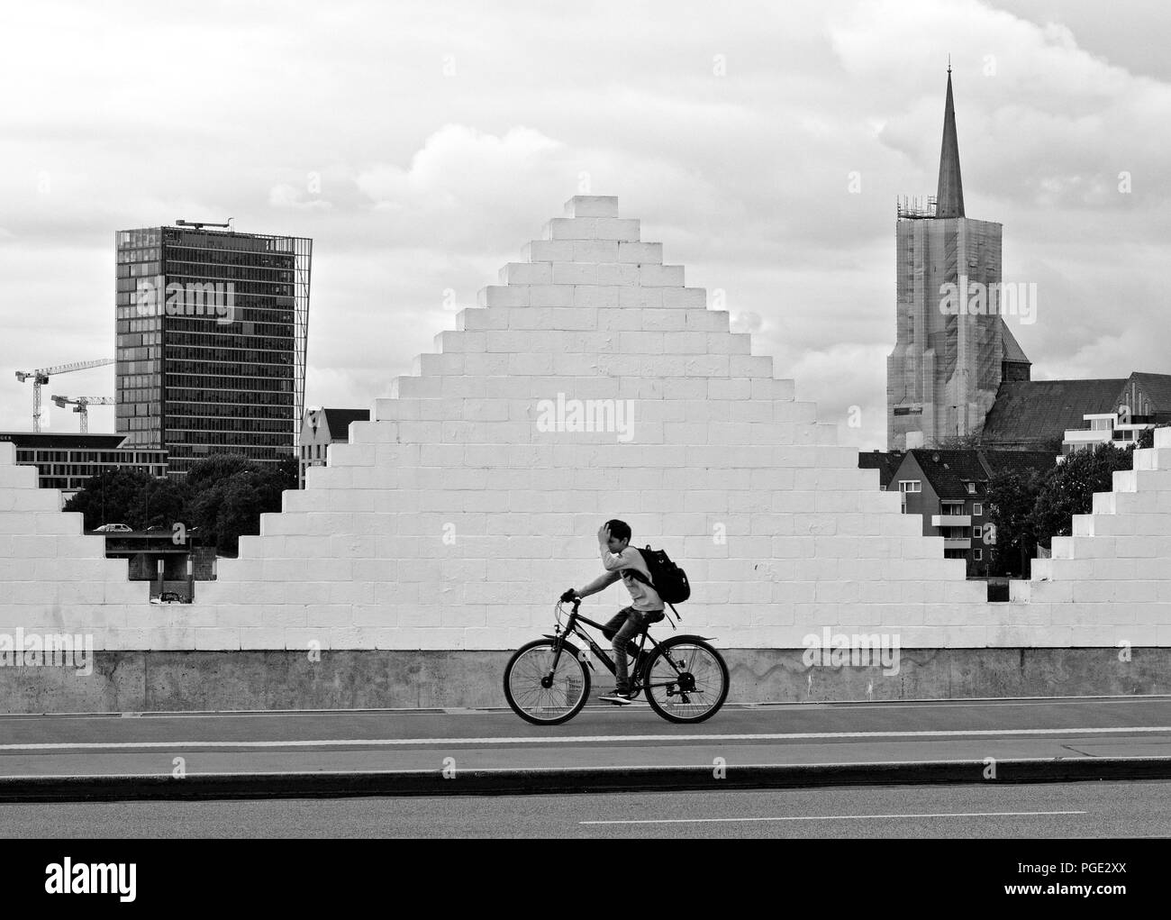 Bremen, Germania - Agosto 14th, 2018 - Un ragazzo corse la sua bicicletta sul marciapiede mentre passa un triangolo bianco a forma di parete di mattoni incorniciata da una chiesa e Foto Stock