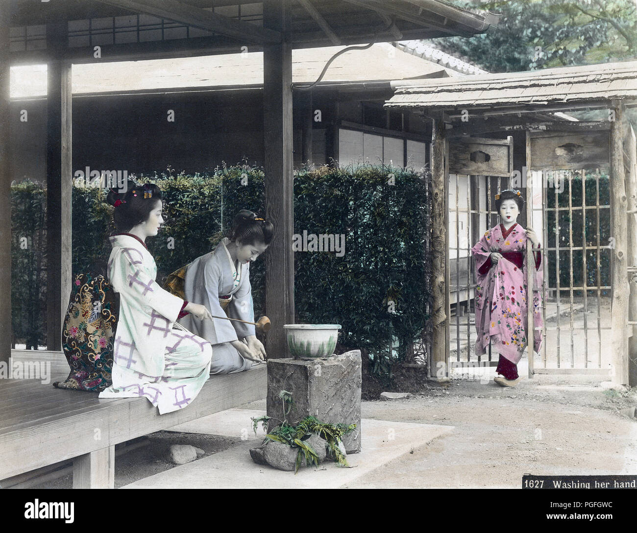[ C. 1890 Giappone - Lavaggio mani ] - una donna in kimono utilizza un Hishaku (siviera) per spruzzare acqua sulle mani di un'altra donna che sta lavando le mani. Le due donne sono seduti sulla veranda di una casa privata. Un terzo donna entra nel giardino attraverso il cancello. Lavaggio delle mani (Temizu) è un importante rituale di purificazione durante l'inserimento di sacrario scintoista motivi, ma è stato anche fatto al momento di entrare in una casa privata. Xix secolo albume vintage fotografia. Foto Stock