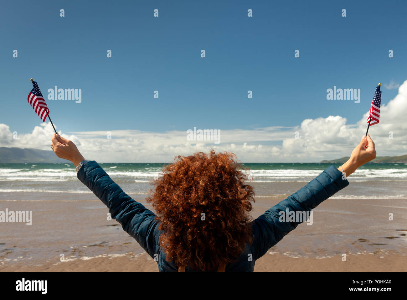 Vista posteriore della giovane donna con il rosso dei capelli ricci rivolta verso il mare mentre si tiene su bandierine americane su di una spiaggia di sabbia sul ventoso giornata soleggiata con cieli blu. Foto Stock