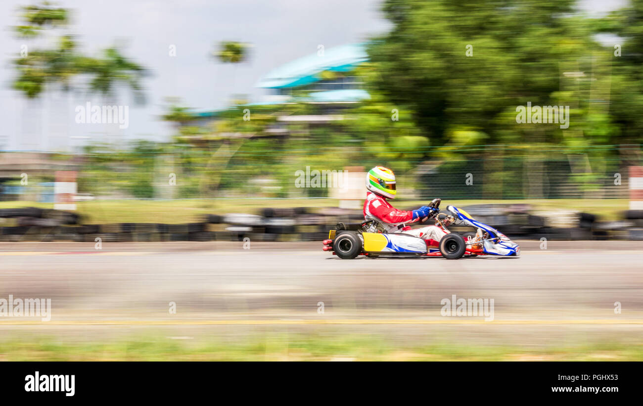 Un corridore professionista con tuta racing nel suo go-kart gareggiando ad City kart racing via sotto un clima caldo, girato con una panoramica con la telecamera verso Foto Stock