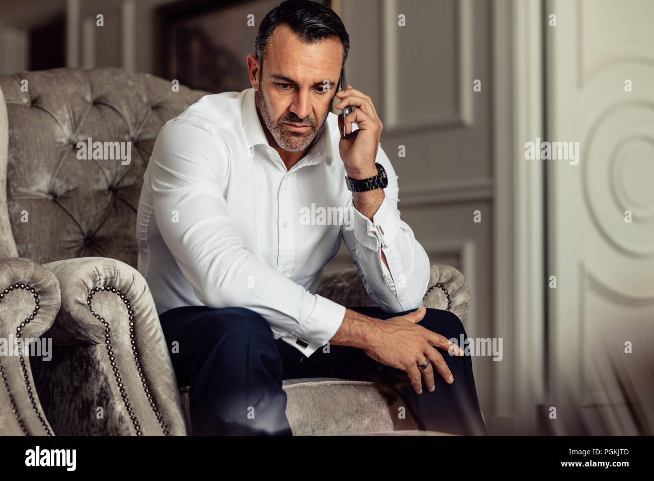 Imprenditore facendo una telefonata dopo essere arrivati nella stanza dell'hotel. Uomo in abbigliamento formale seduti su una sedia e parlare sullo smartphone. Foto Stock