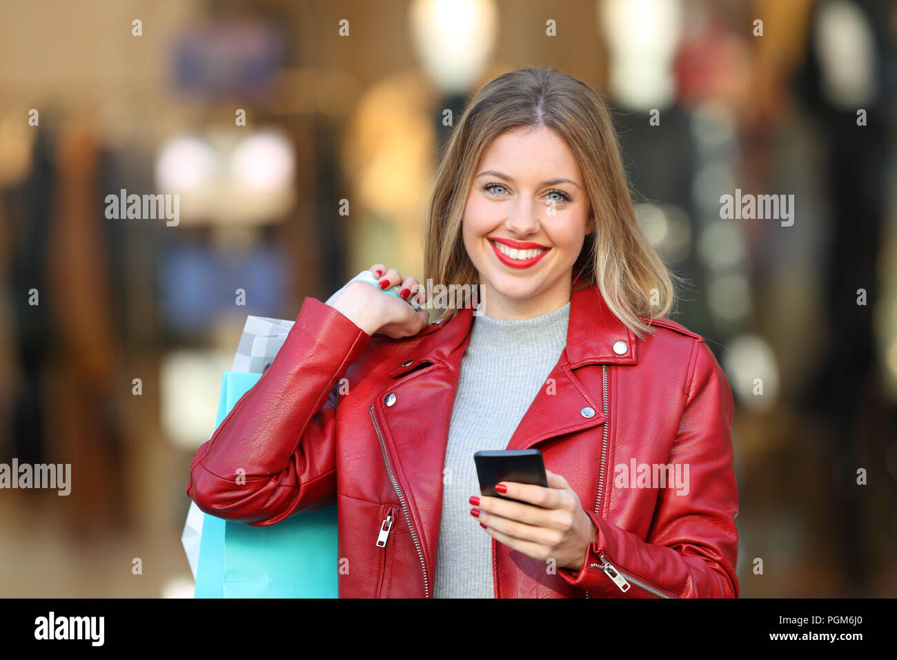 Happy shopper in posa tenendo un telefono intelligente e borse per lo shopping in strada con una vetrina in background Foto Stock
