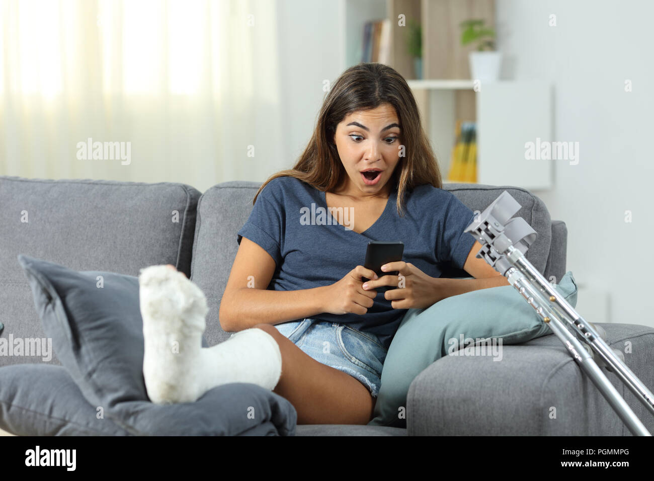 Sorpreso donna disabile la lettura del contenuto del telefono seduta su un divano nel salotto di casa Foto Stock