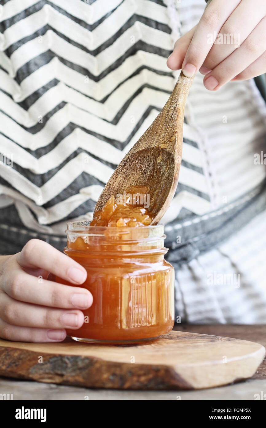 Giovane donna con le mani in mano per mettere un cucchiaino di marmellata di melone in un vaso come parte di un processo di inscatolamento il frutto dal suo giardino. Estrema profondità o Foto Stock