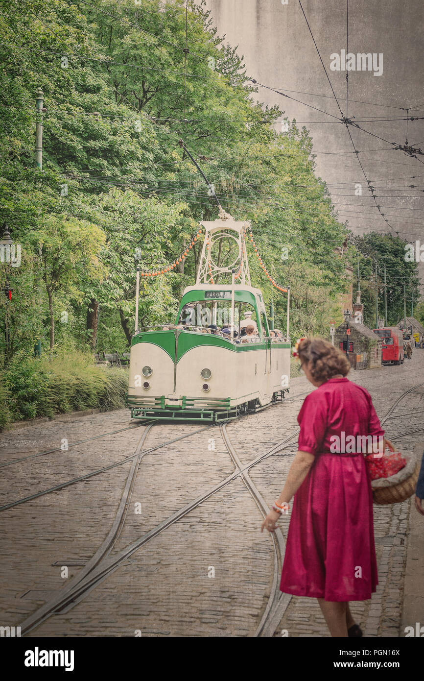 Crich tramvia Village, 1940s weekend. Età-look vintage street scene con open-top tram viaggiano in strada di ciottoli. Signora in abito rosso attende a croce. Foto Stock