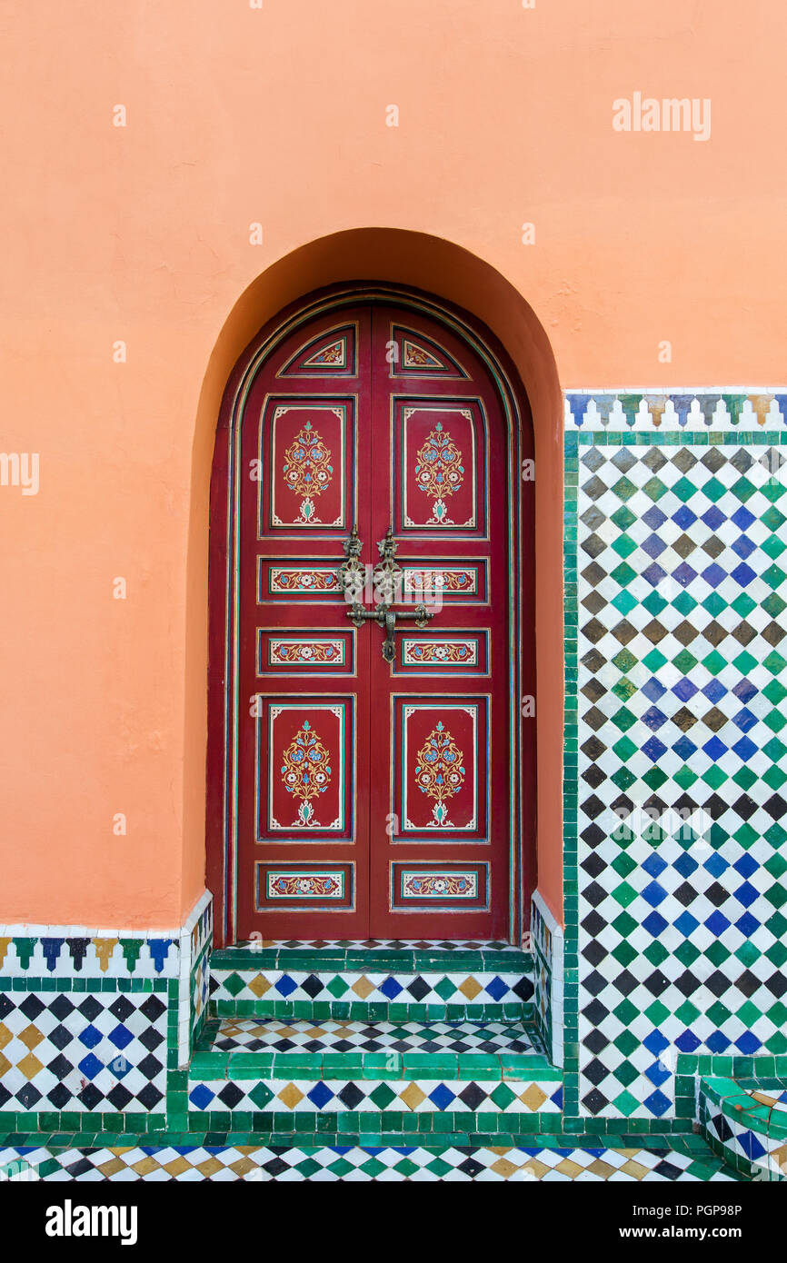 Marocchino esterno arcuato porta, verniciato di rosso con disegni floreali, inset in un arancione intonaco decorato con piastrelle a mosaico. Marrakech, Marocco Foto Stock