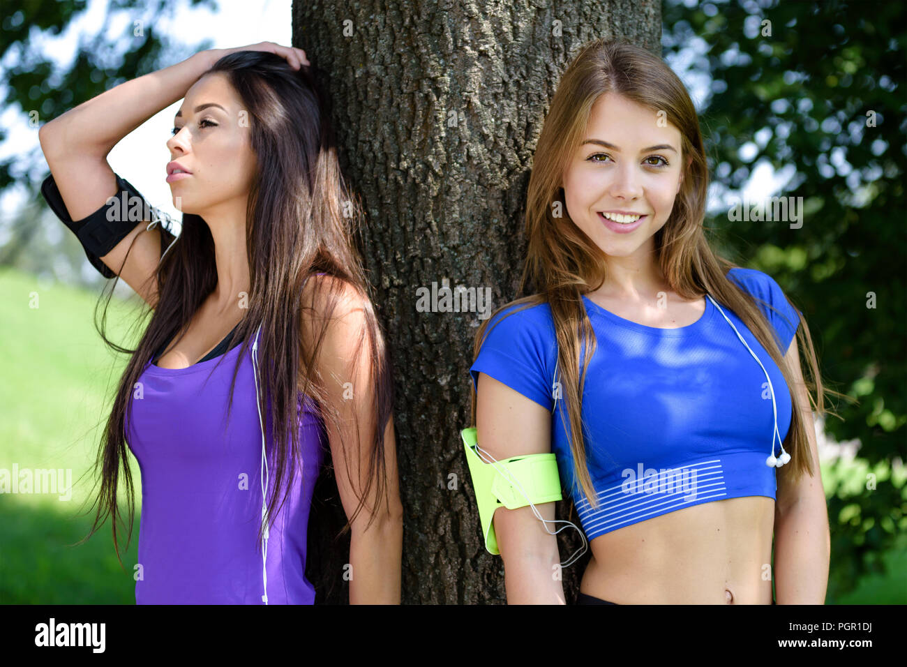 Ritratto di due belle e giovani ragazze che riposa in un parco appoggiata contro un albero dopo un allenamento in una giornata di sole Foto Stock
