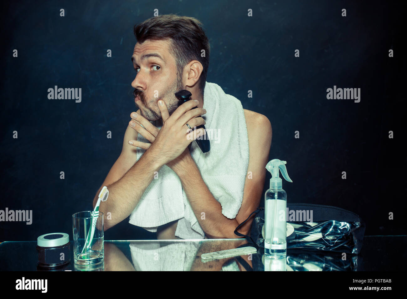 Il giovane uomo in camera da letto seduto di fronte allo specchio di graffiare la sua barba a casa. Le emozioni umane e il concetto di stile di vita Foto Stock