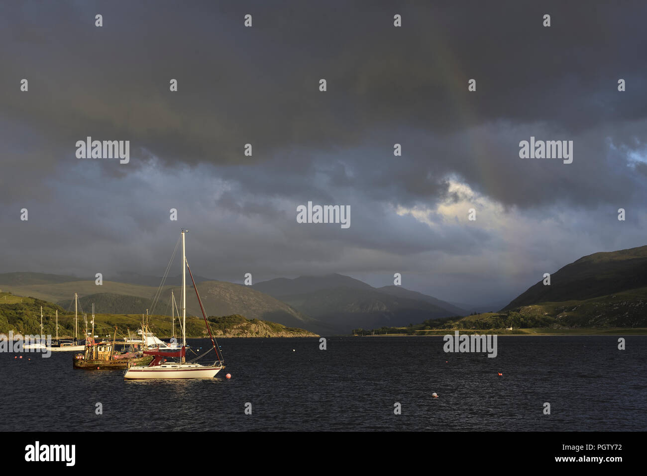 Il lungomare a Ullapool, nel nord della Scozia, con barche, nubi e un debole rainbow. In lontananza sono le montagne Fannich. Foto Stock