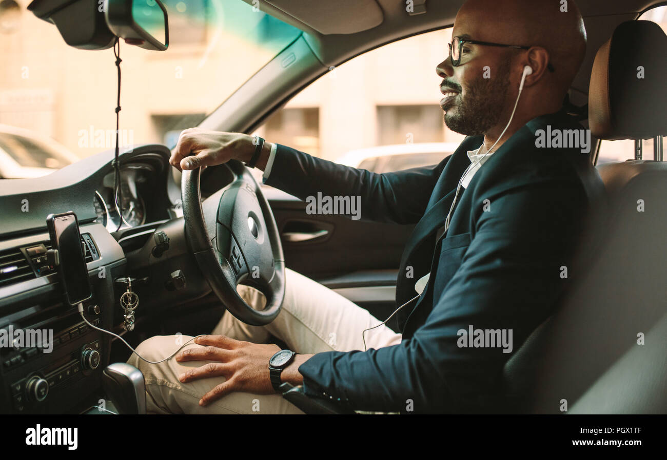 Vista laterale del giovane uomo in tuta ascoltare musica mentre si guida in auto. Imprenditore africano alla guida di un veicolo per l'ufficio. Foto Stock