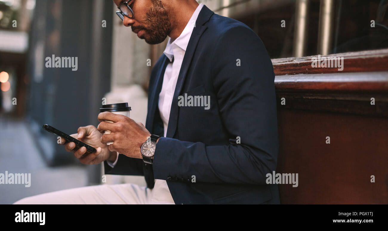 Imprenditore africano seduto fuori con caffè e utilizzando il telefono cellulare. Uomo in completo relax all'aperto mediante il texting su strada sul suo cellulare e drinkin Foto Stock
