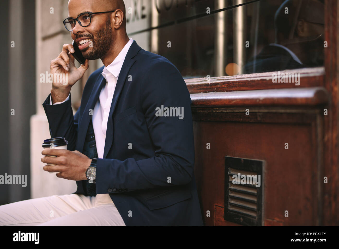 Felice l'uomo africano di relax all'aperto con caffè e parlando al telefono cellulare. Giovane uomo nel business suit seduto fuori e parlare al telefono. Foto Stock