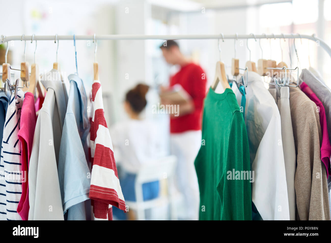Riga di casual t-shirt e altri vestiti appesi su rack con due giovani stilisti lavorano nelle vicinanze Foto Stock