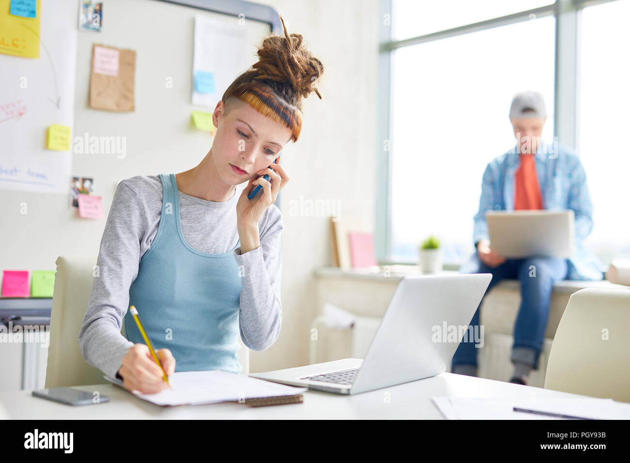 Grave concentrata hipster ragazza con i timori parlando al telefono cellulare e a rendere note in blocco schizzi mentre si lavora in un ufficio moderno Foto Stock