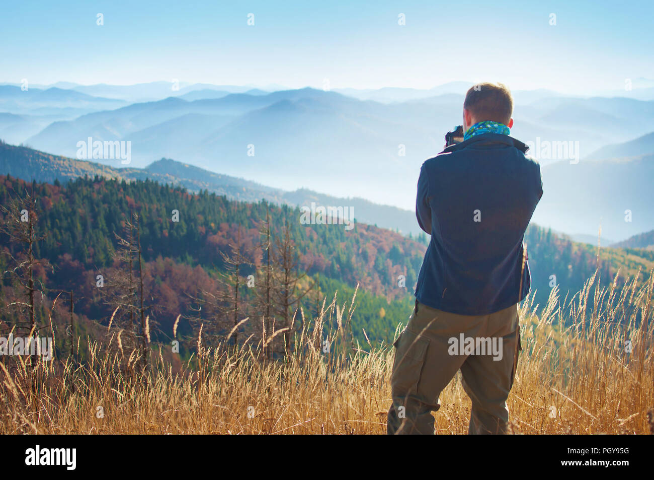 Un giovane uomo in piedi con la schiena rivolta, tenere la fotocamera e fare foto di una catena montuosa ricoperta in rosso, arancione, giallo bosco di latifoglie unde Foto Stock