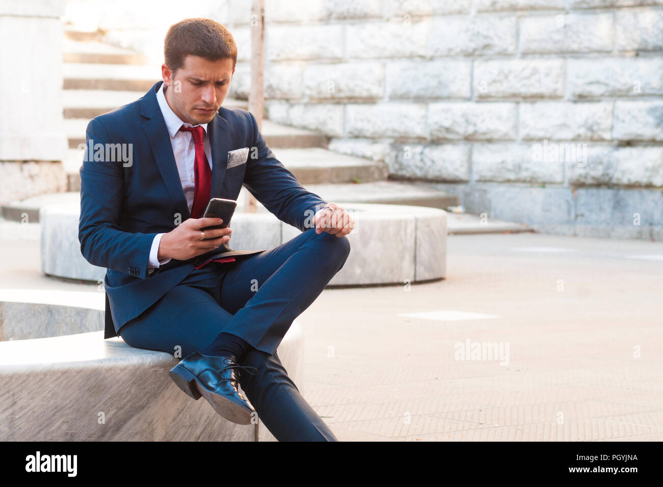 Giovane imprenditore bello in un vestito e un tirante consulta lo smartphone seduto su un banco di marmo nel parco Foto Stock