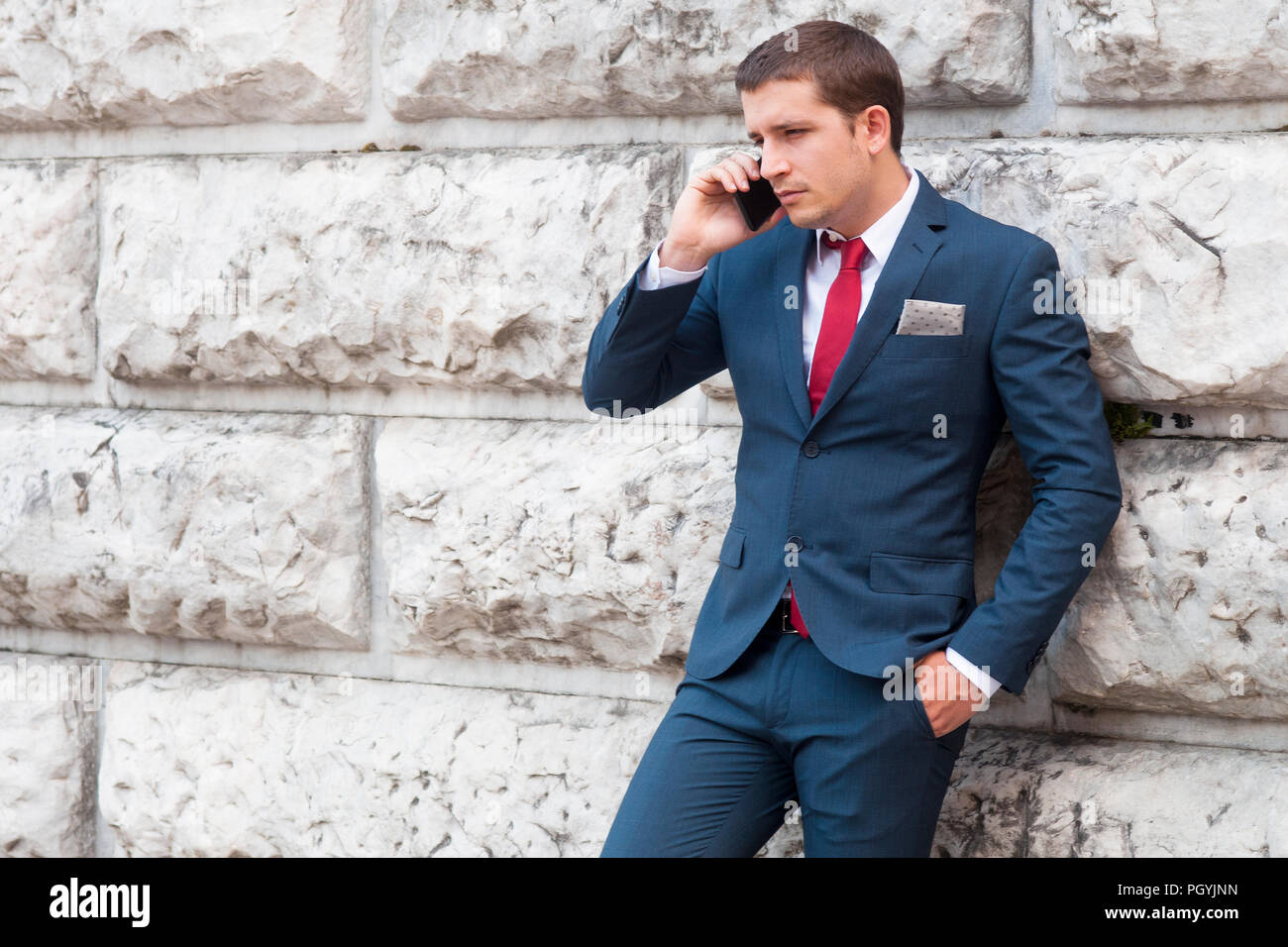 Giovane imprenditore bello in un vestito e un tirante parlando a smartphone contro una parete di marmo Foto Stock