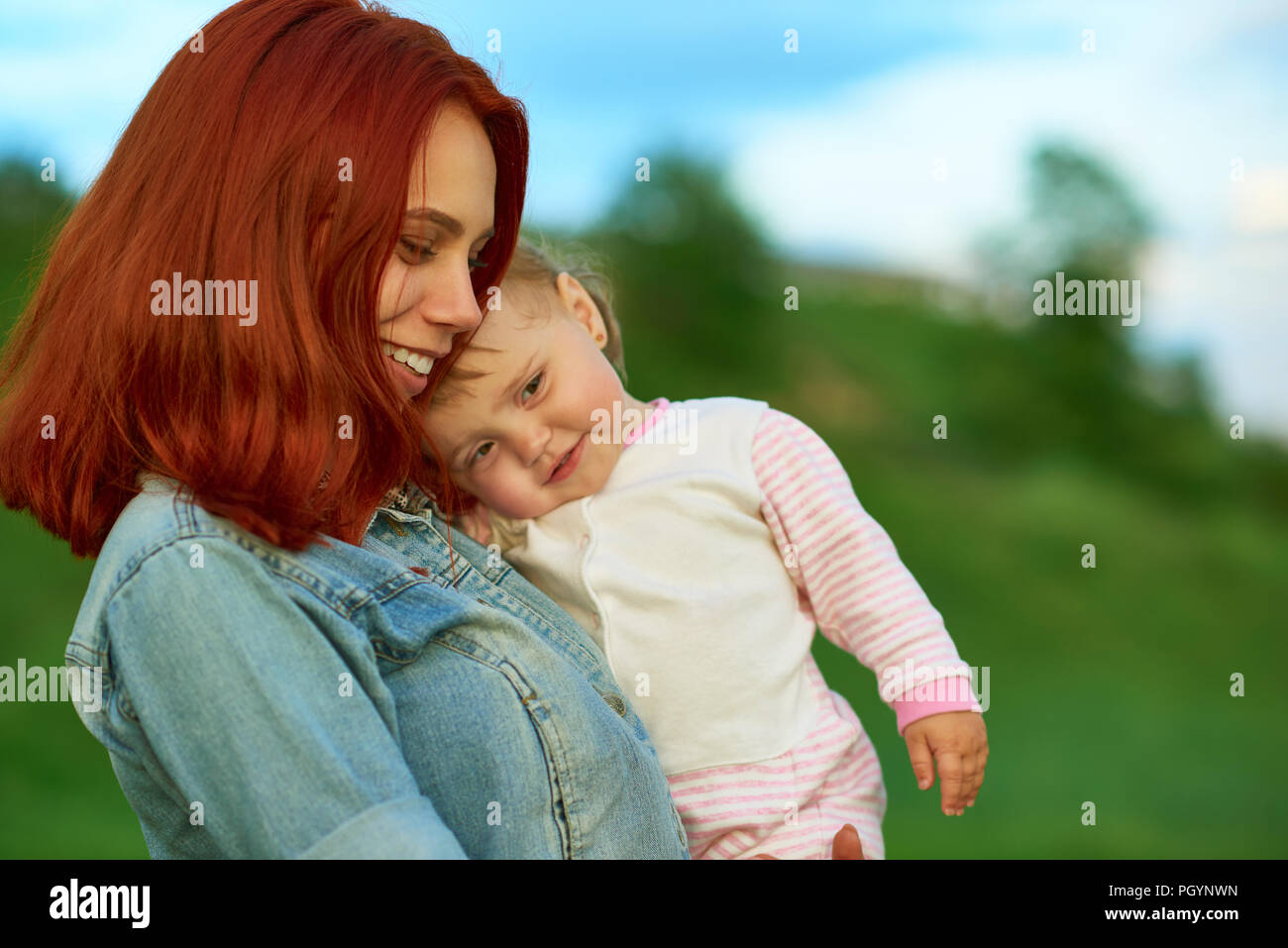 Vista laterale della madre mantenendo grazioso fanciullo in posa sul campo verde. Mom abbracciando bambino felice, con splendide caratteristiche della faccia, capelli rossi, indossando blue je Foto Stock