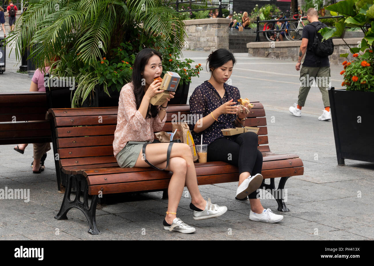 Due giovani donne asiatiche seduta su una panchina e mangiare hamburger da asporto nel luogo Jacques-Cartier nella Città Vecchia, Montreal, QC, Canada Foto Stock