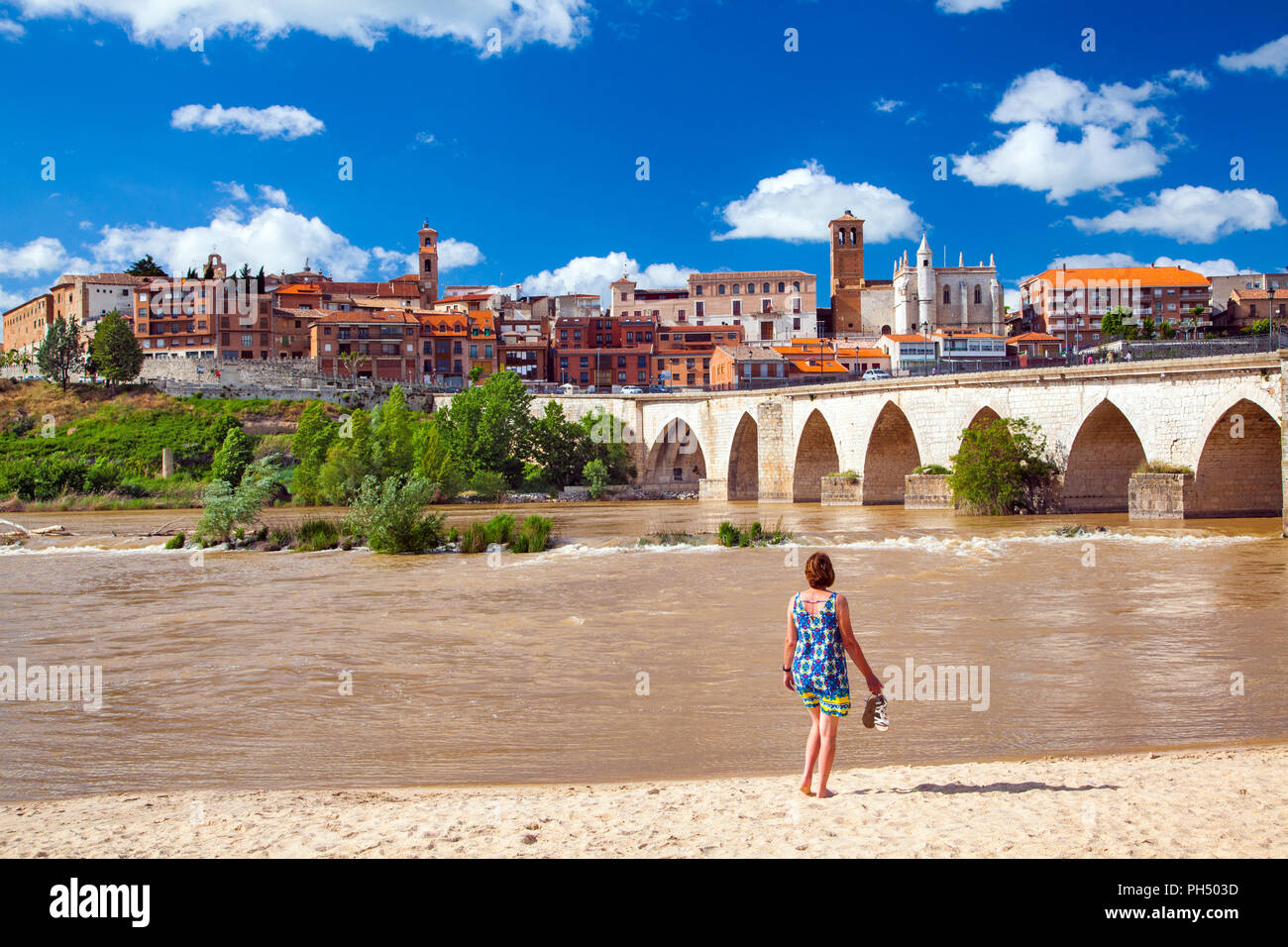 La donna a piedi nudi azienda scarpe sulle spiagge artificiali sul fiume Douro con una vista della città medievale di Tordesillas in distanza Spagna Foto Stock