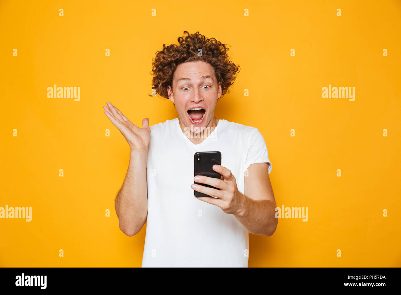 Emozionato uomo europeo in casuale t-shirt bianco urlando mentre guardando smartphone isolate su sfondo giallo Foto Stock