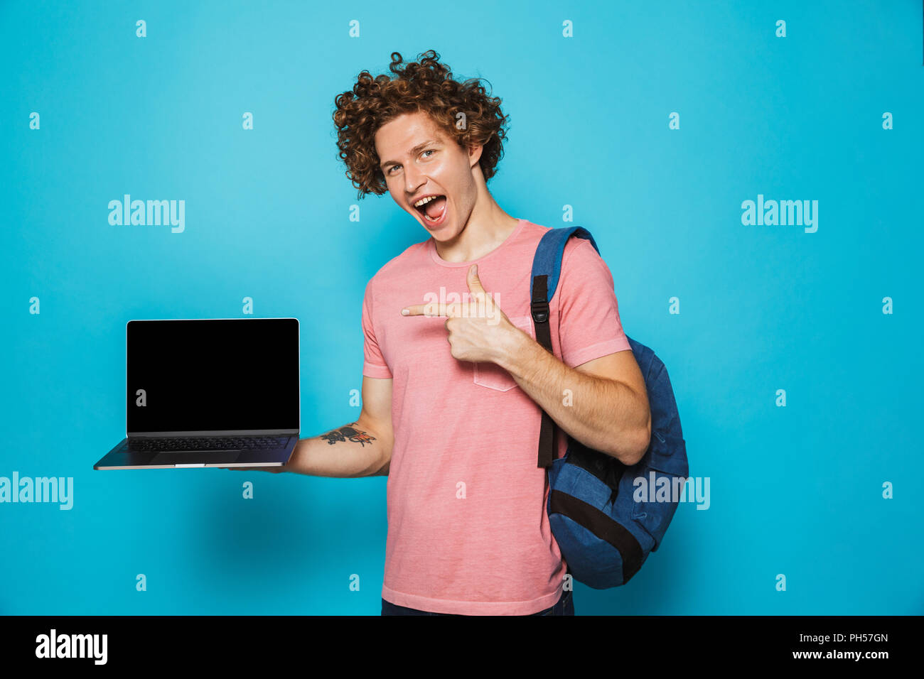 Foto di università ragazzo con i capelli ricci di indossare un abbigliamento informale e uno zaino tenendo aperto il laptop e puntare il dito a schermo isolate su sfondo blu Foto Stock