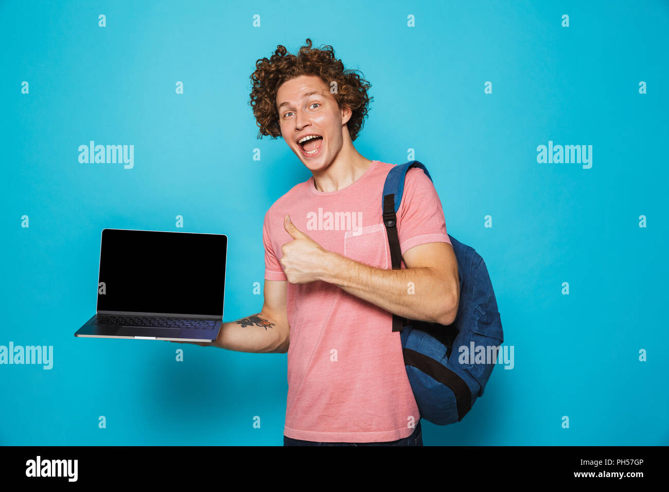 Foto di università ragazzo con i capelli ricci di indossare un abbigliamento informale e uno zaino tenendo aperto il laptop e mostrando il pollice in alto isolato su sfondo blu Foto Stock