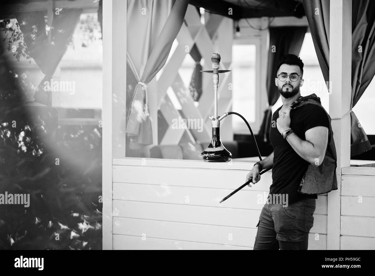 Barba elegante uomo arabo in bicchieri e t-shirt nera di fumare narghilè all'aperto. Modello arabo avente il resto. Foto Stock
