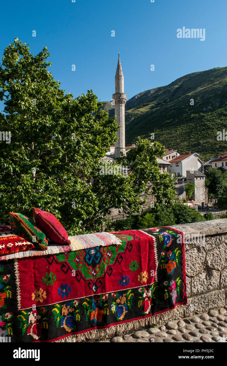 La Bosnia Erzegovina, Europa: lo skyline della città di Mostar con cuscini e tappeti decorati su un muro di pietra e il minareto della Nezir agina moschea dzamija Foto Stock
